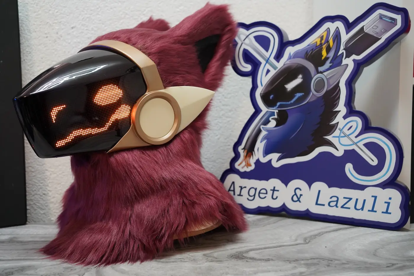 Burgundy Protogen head with small round orange eyes and a wide grin on the LED visor