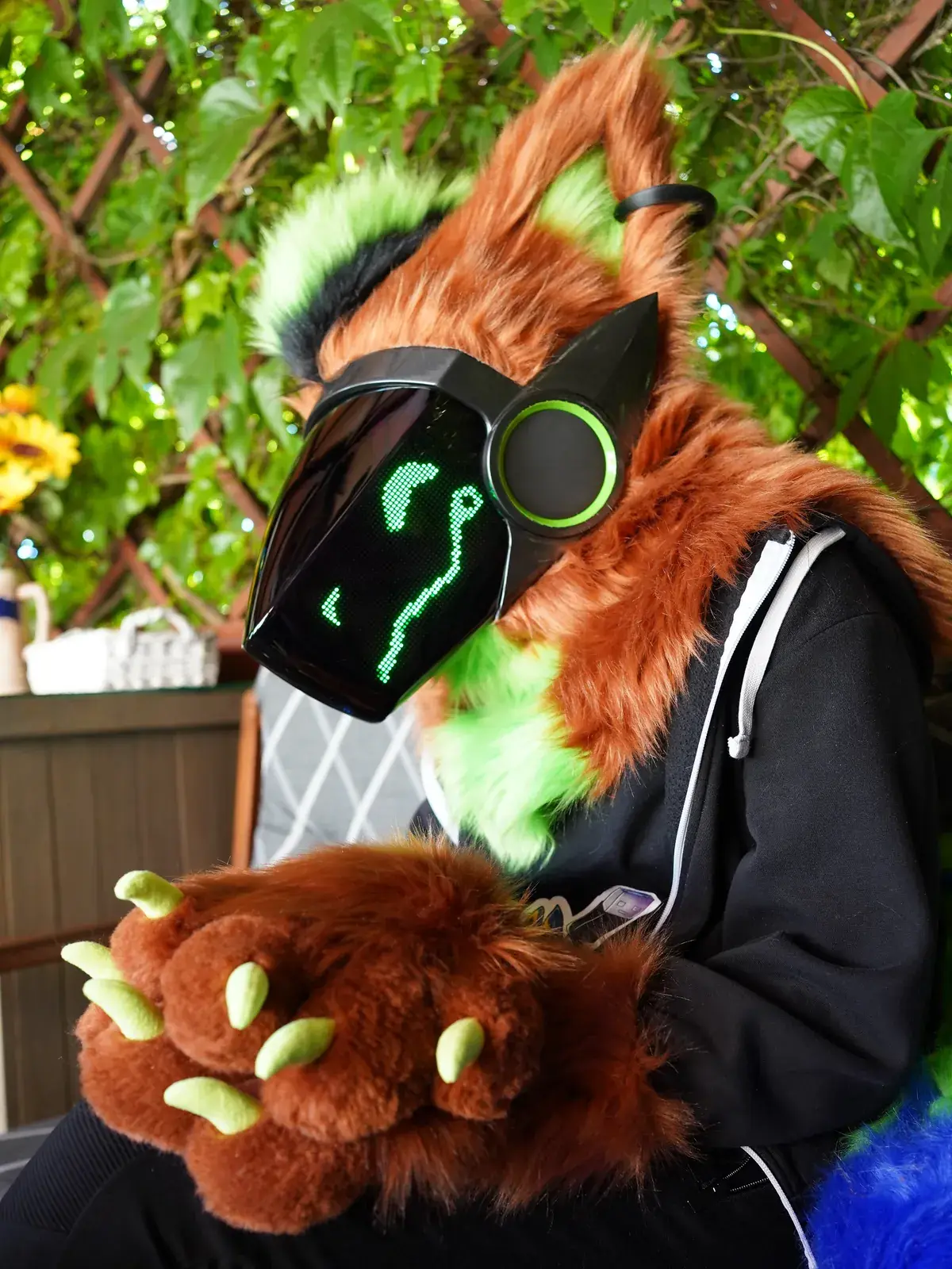 Rust brown Protogen partial under a leafy pergola, green pixel face on the LED visor and hand paws with green claws