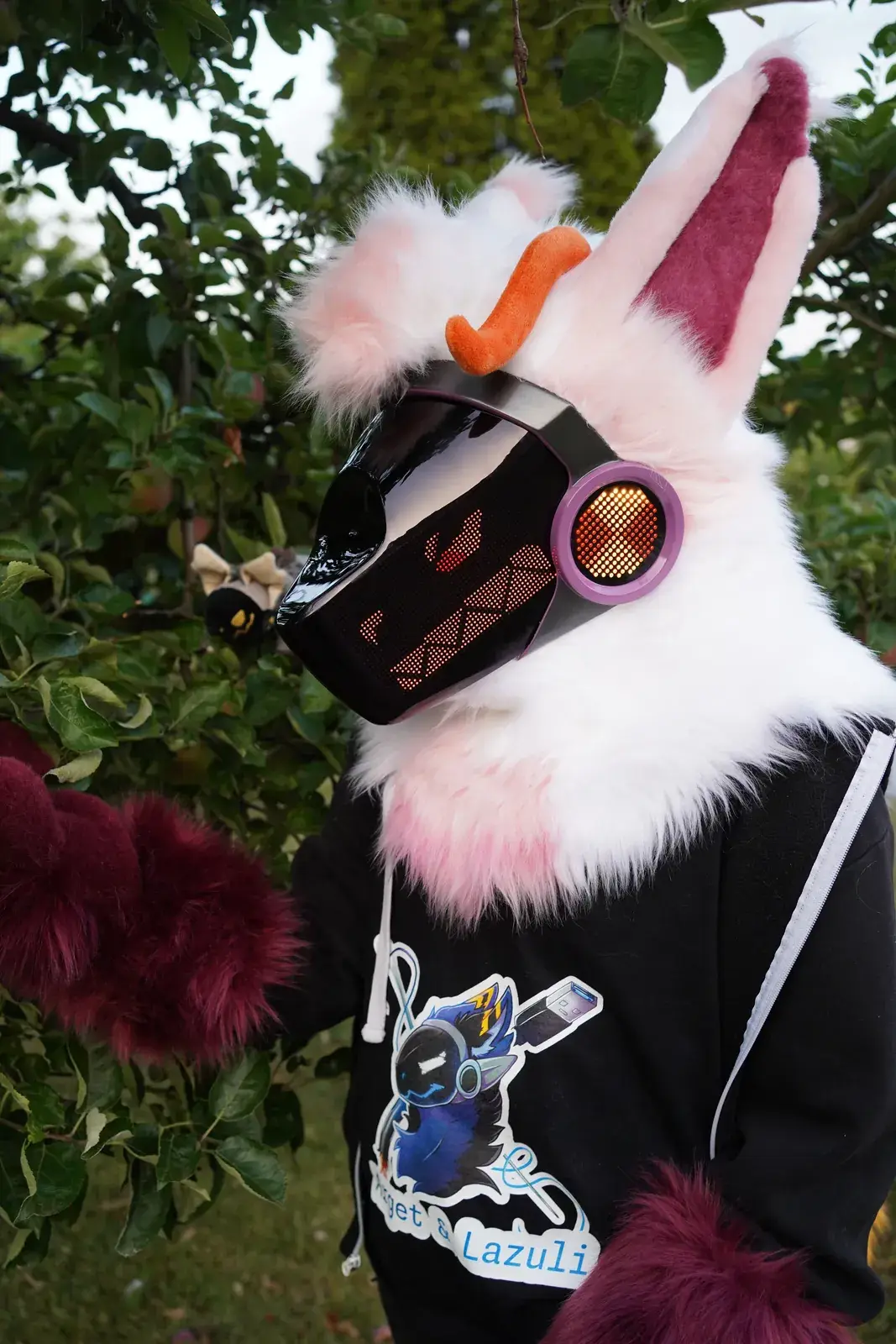 Close up of a white and pink Protogen mini partial in an apple tree with a small figurine, red grin with teeth on the LED visor