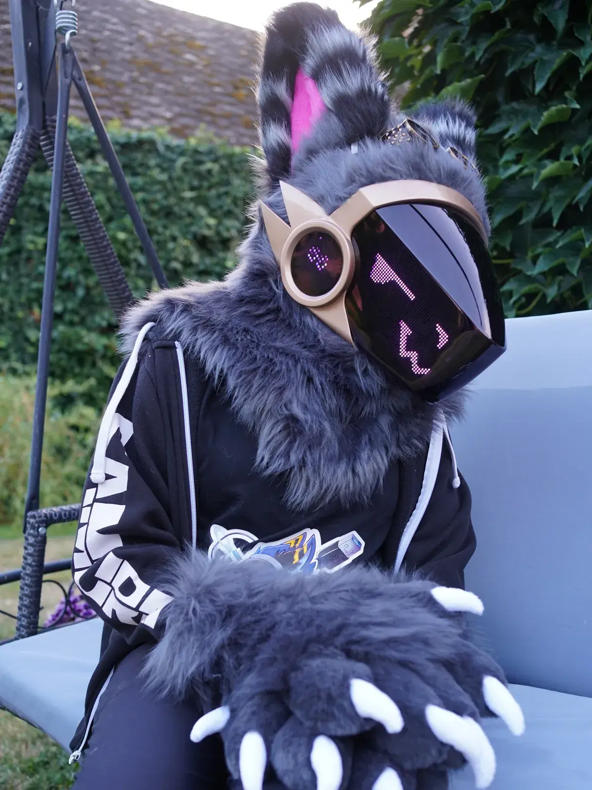 Grey Protogen full partial sitting on a garden swing, grey hand paws with white claws and a purple face on the LED visor