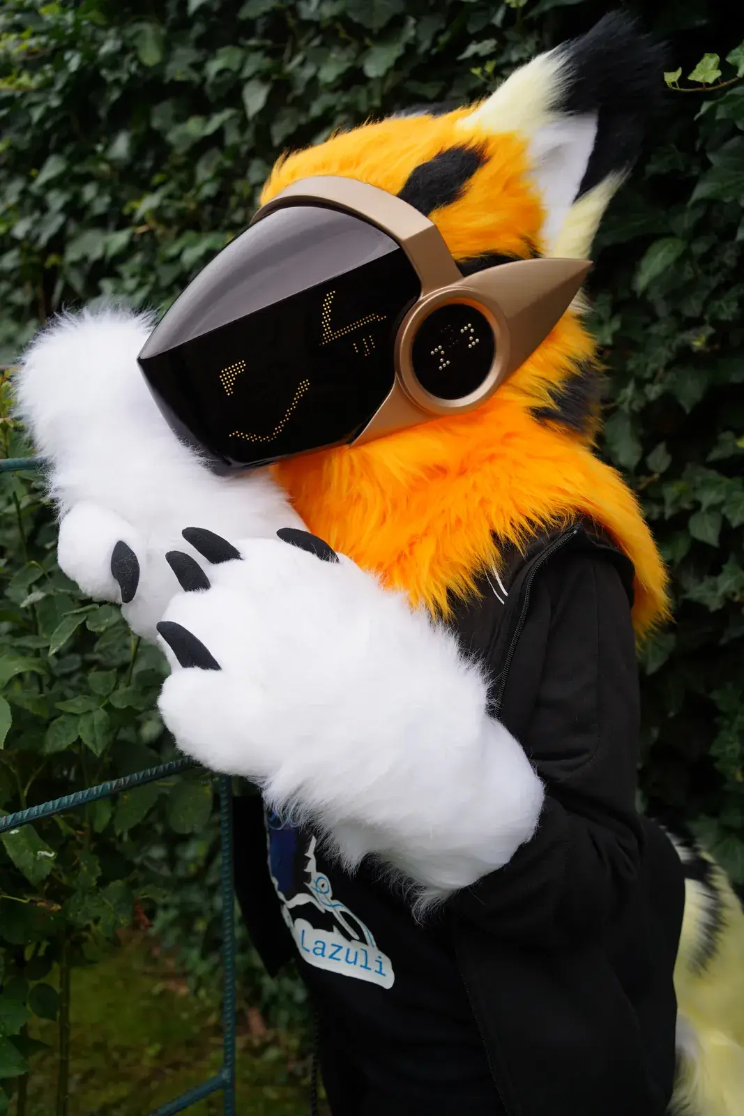 Orange Protogen mini partial by an ivy wall with a white hand paw raised