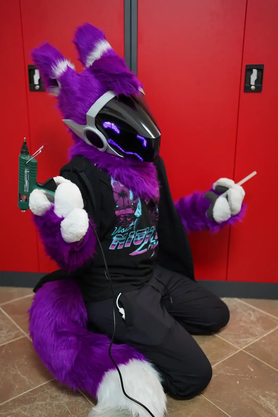 Purple fursuit partial seated on the floor holding a glue gun, black LED pixel visor showing blue eyes