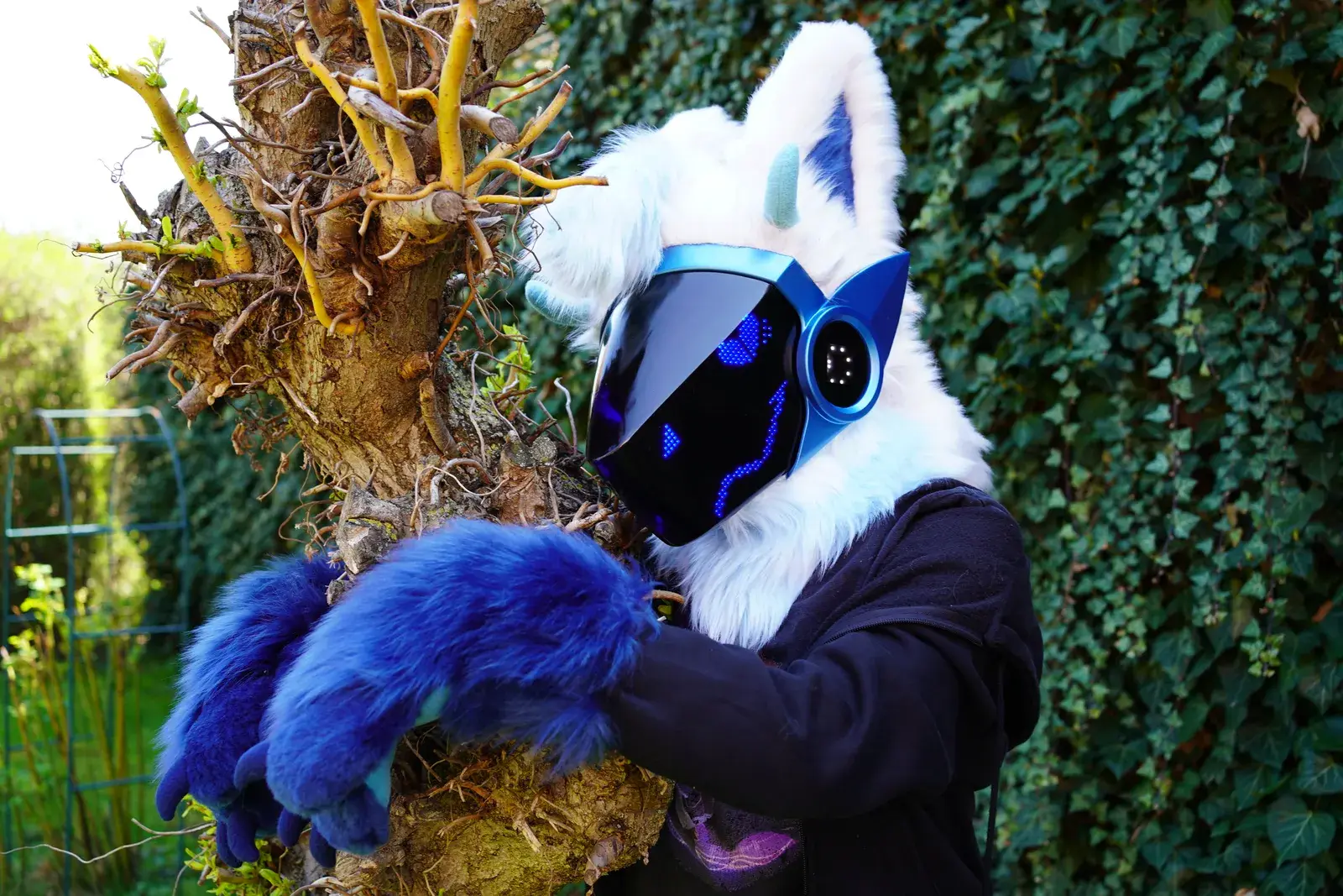 White and blue Protogen partial with a dark visor showing blue pixel eyes, hugging a tree outdoors