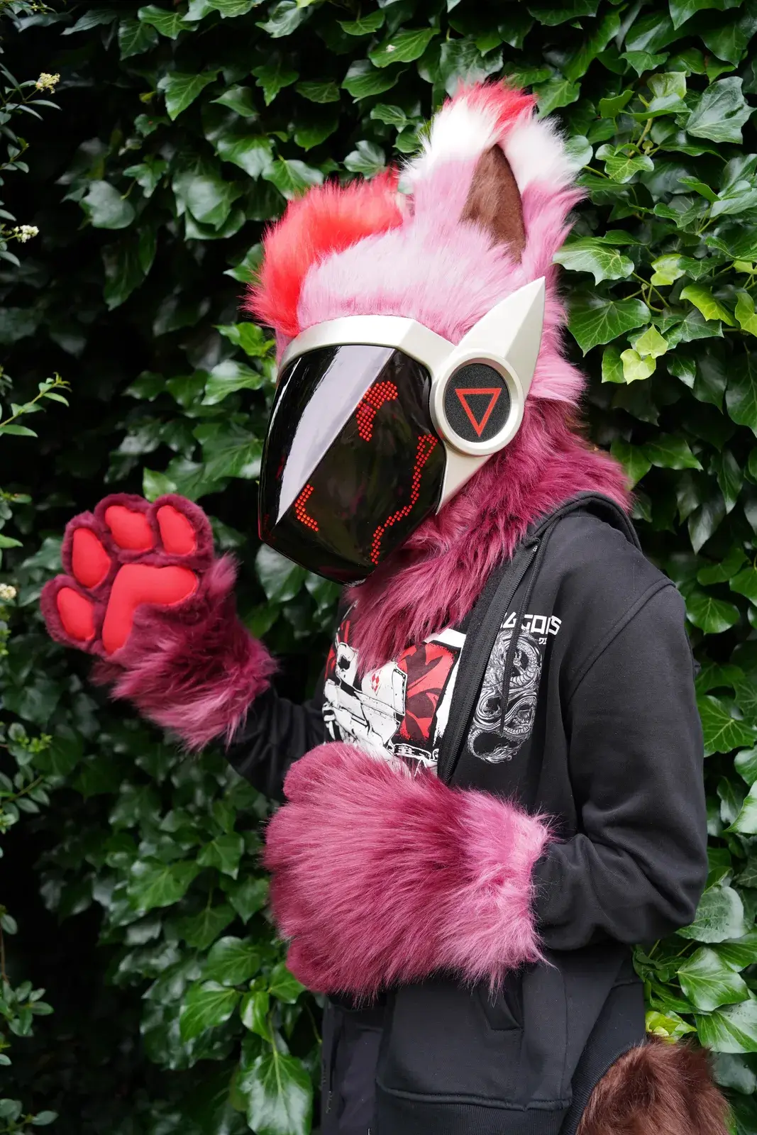 Pink and magenta Protogen partial with a red pixel visor, waving a fluffy red paw toward the camera