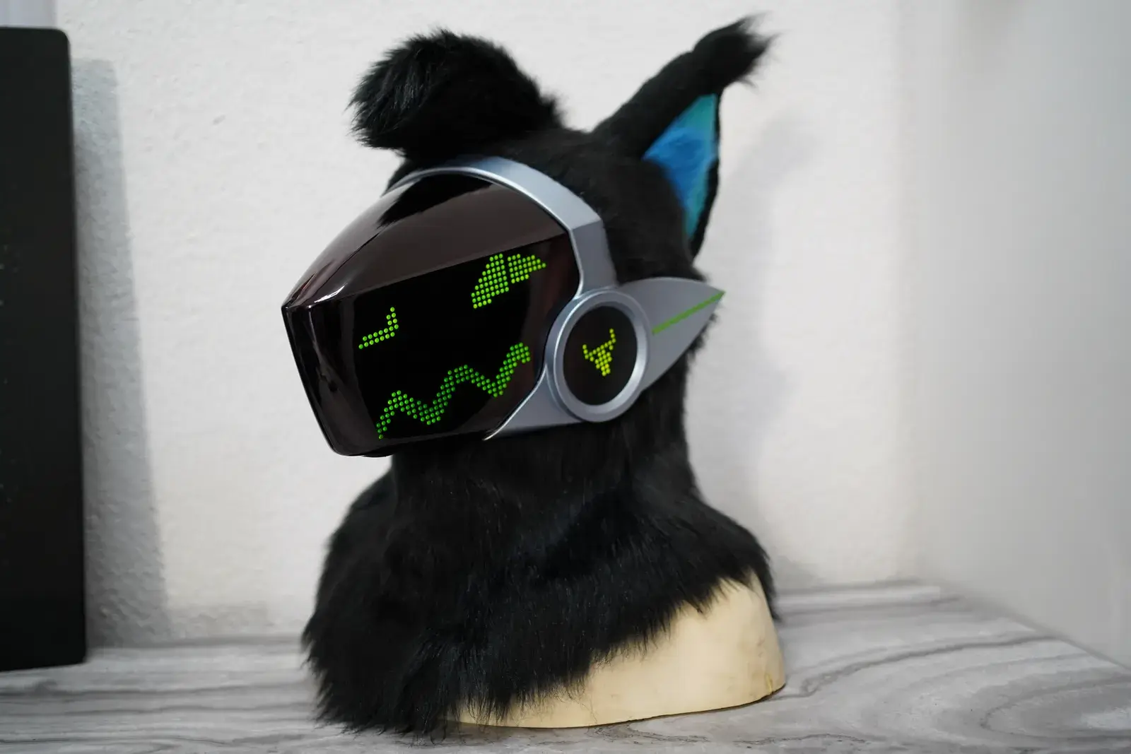 Black Protogen head on a stand with a silver visor displaying green pixel eyes and teal ear interiors