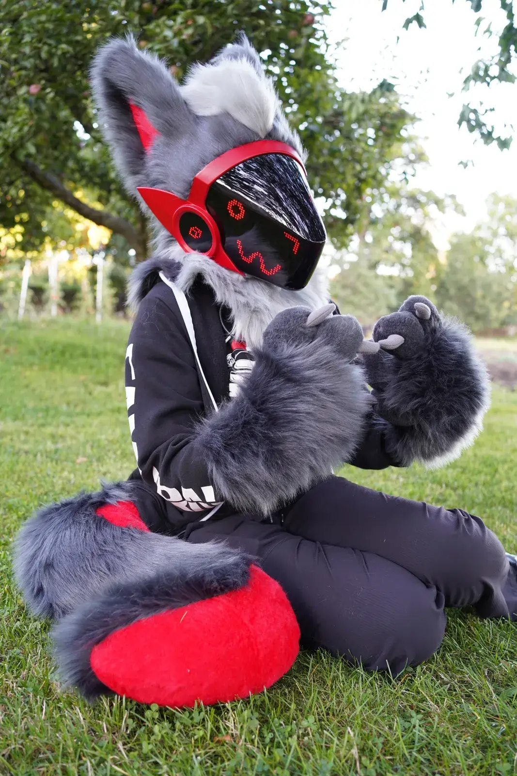 Gray and red Protogen partial sitting on grass, black pixel visor with red trim, fluffy gray paws and red tail