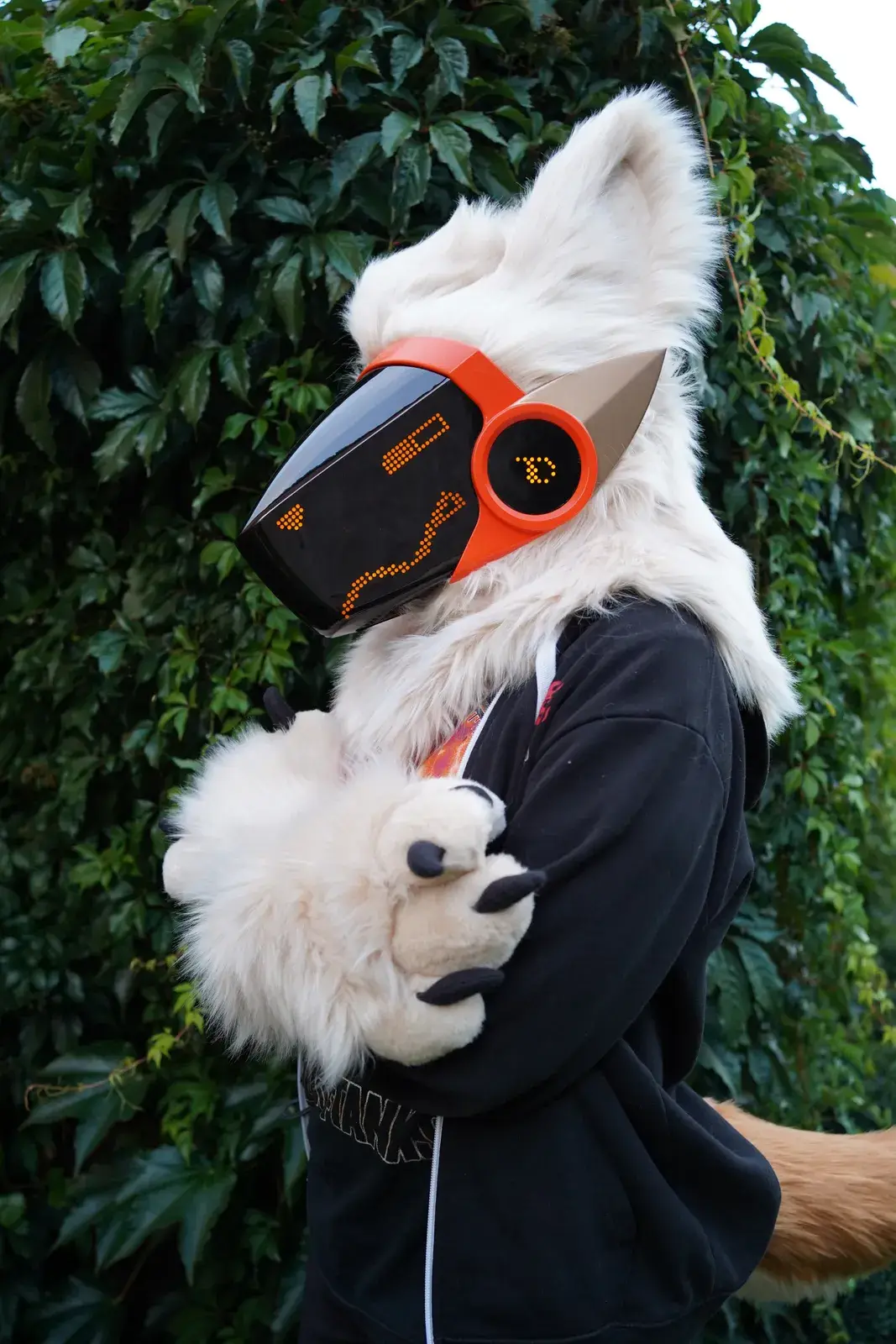 White Protogen partial with an orange trimmed black visor showing amber pixel eyes and fluffy white paws outdoors