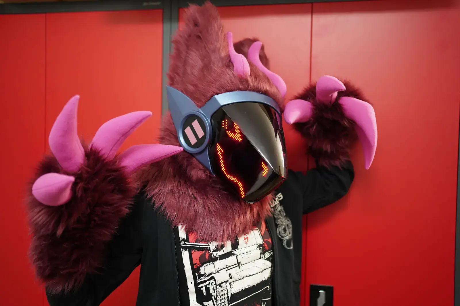 Dark red fursuit partial with many pink horns and a blue gray visor showing red pixel eyes against a red wall