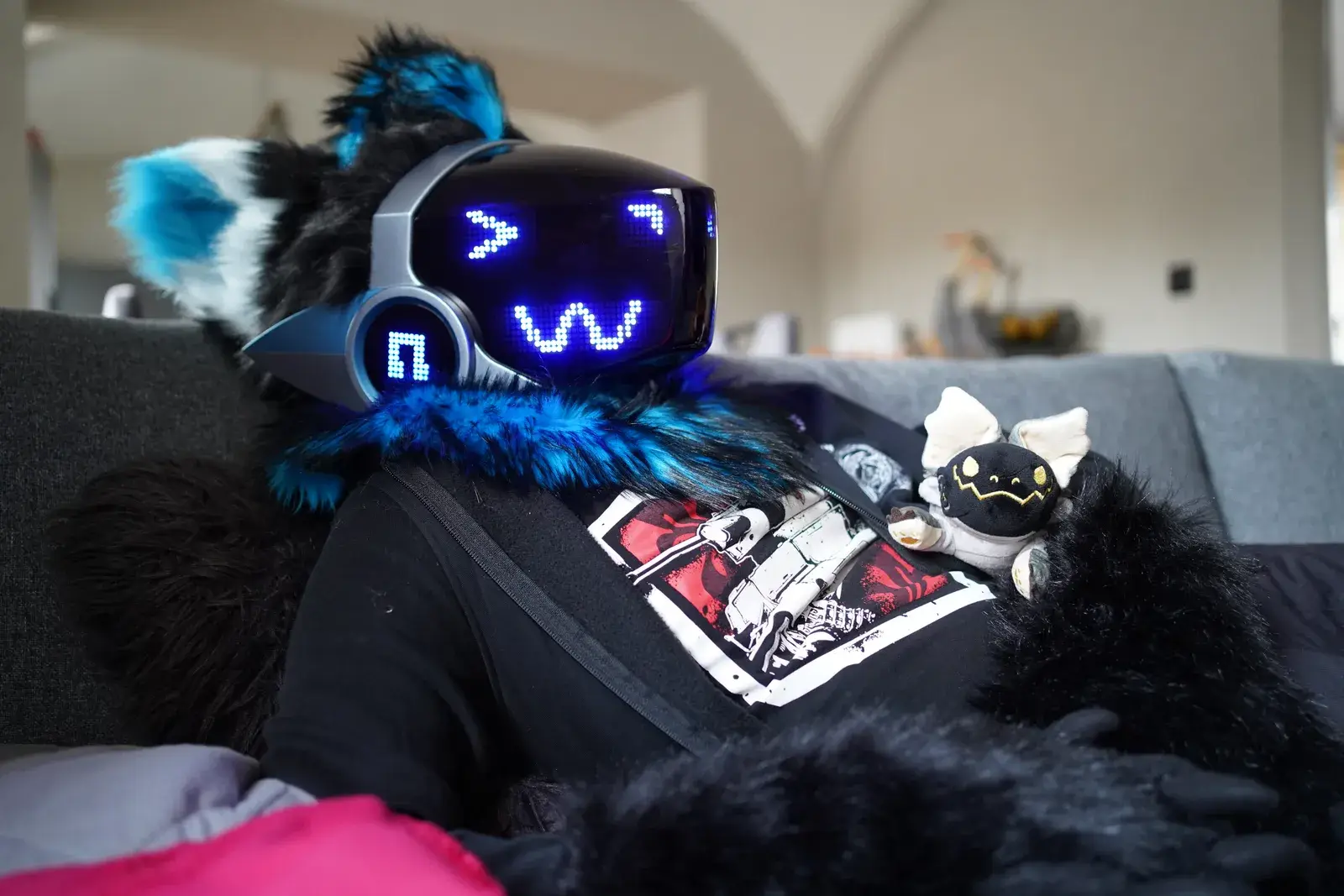 Black and teal Protogen head on a sofa, blue pixel visor with a squiggly eye pattern and teal fluffy ears