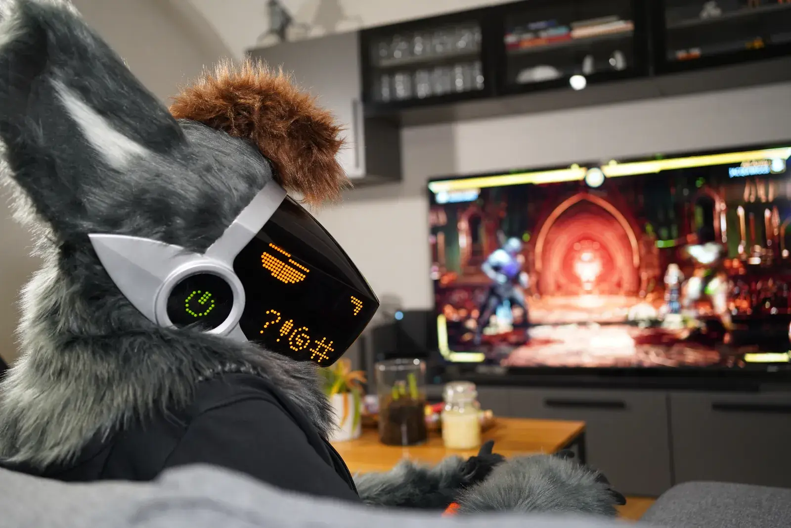 Gray Protogen partial reclining indoors facing a gaming TV, white visor with an amber dot matrix text display