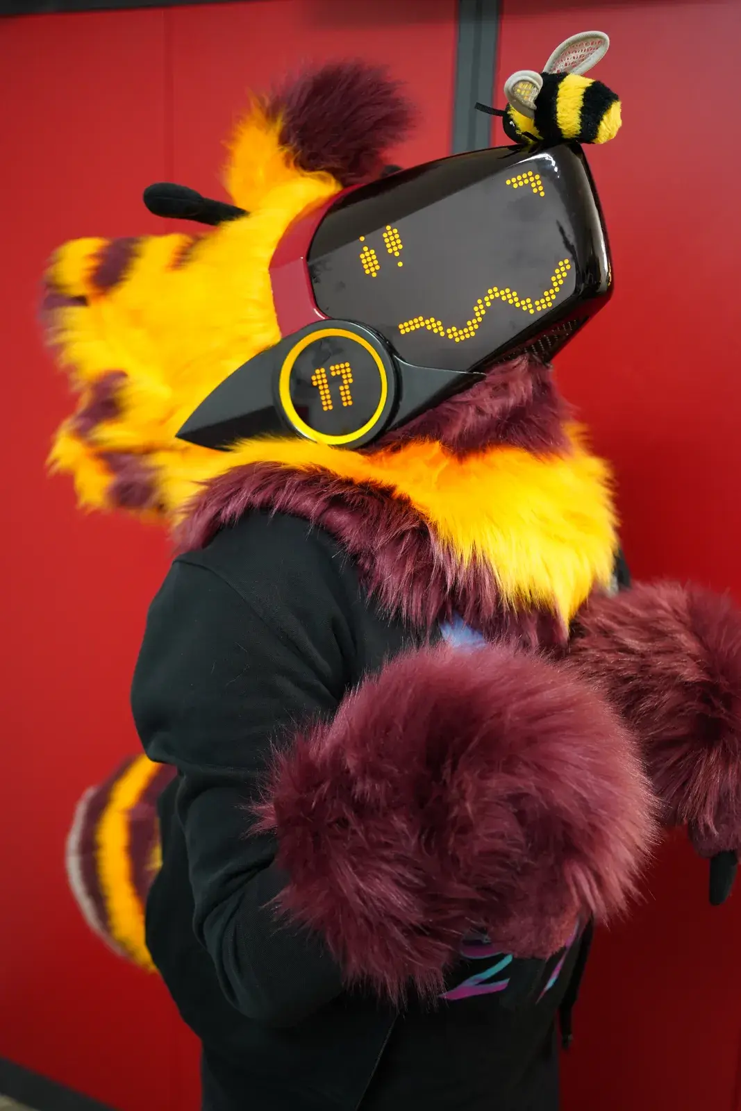 Yellow and dark red Protogen partial in profile, black visor with an amber LED smile and a toy bee on the head
