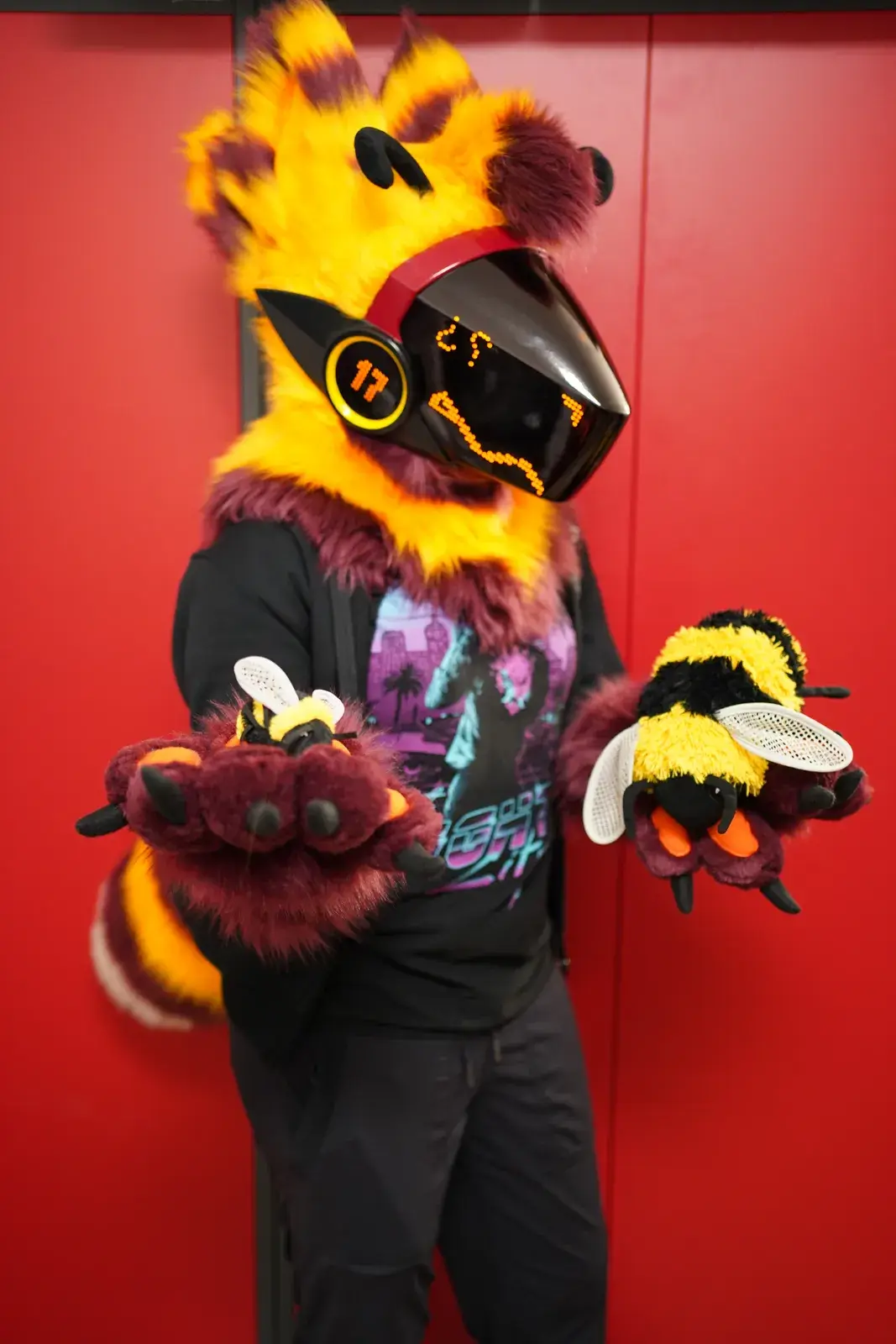 Yellow and dark red Protogen partial holding two bumblebee plush toys, black visor with an amber LED face and dark red paws