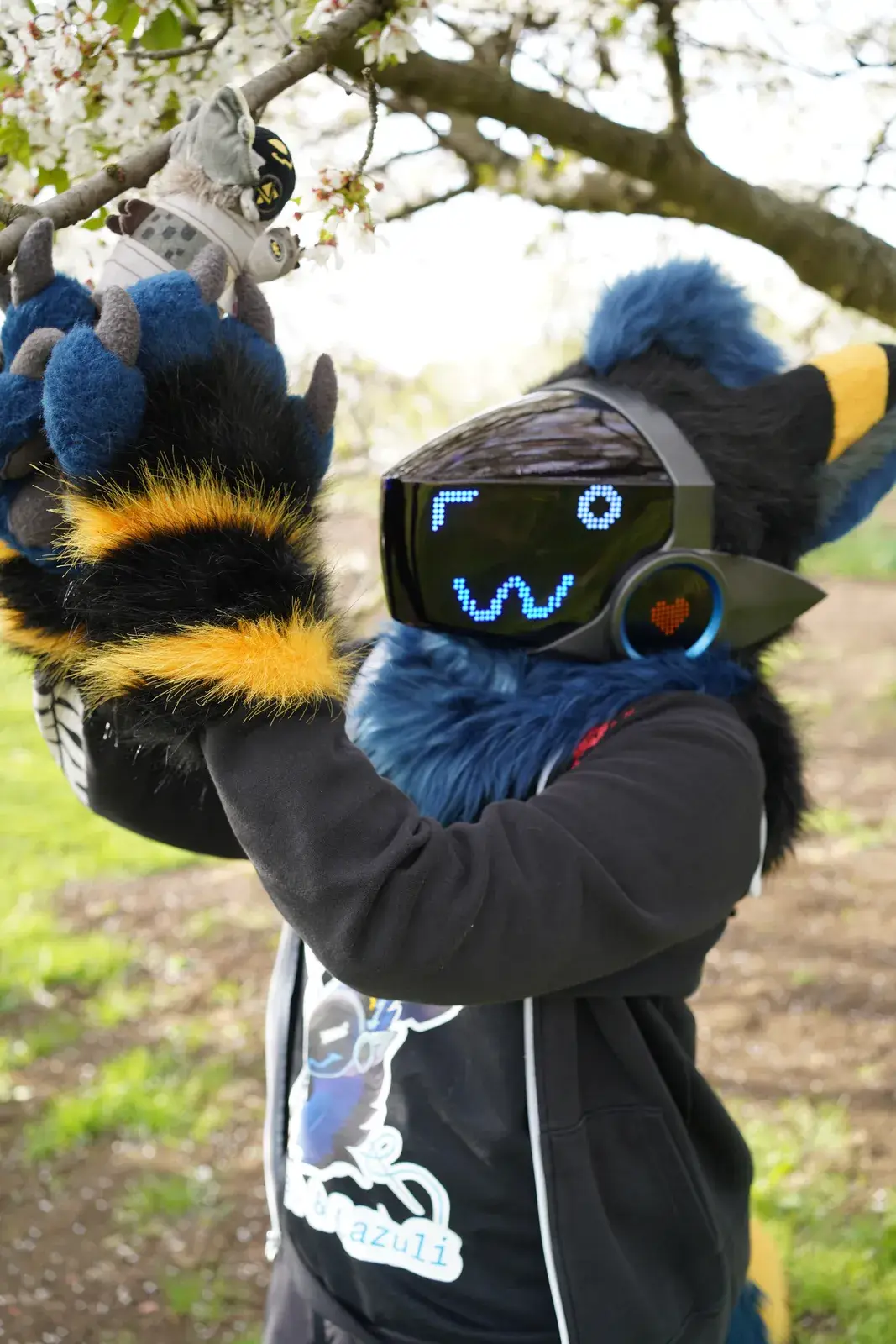 Blue, black and yellow Protogen partial holding a flowering branch at a blossom tree, dark visor with blue pixel eyes