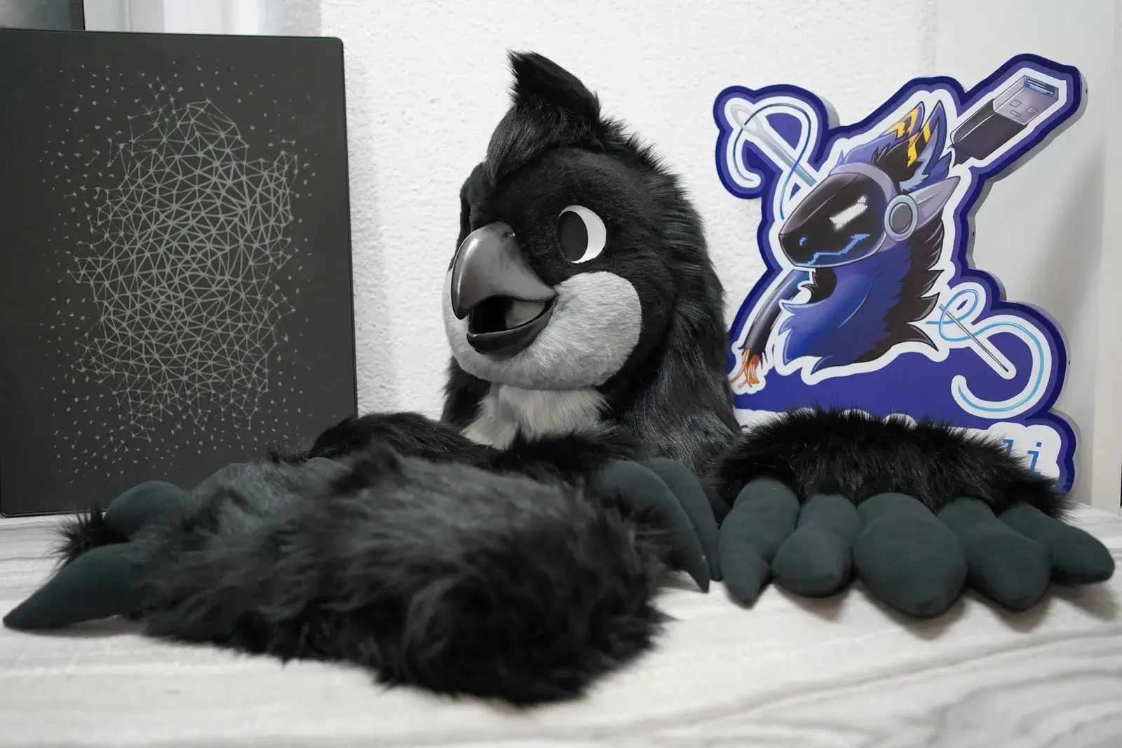 Black bird fursuit head with a gray beak and white eye patches, laid out indoors with black handpaws and a branded standee