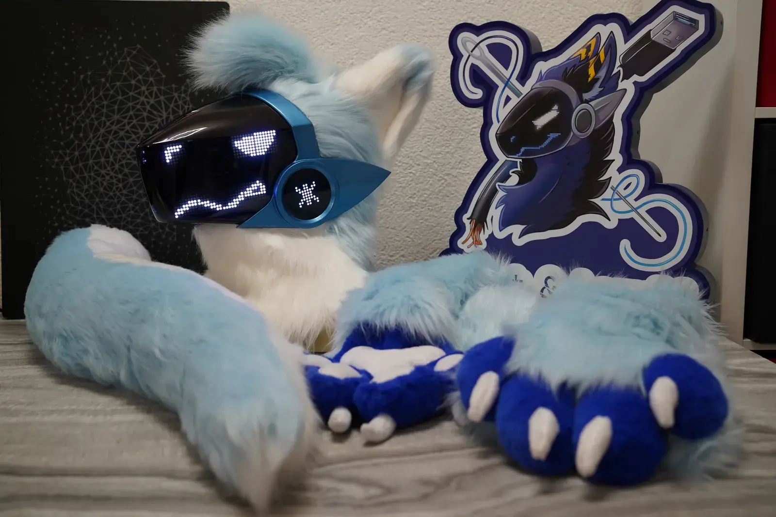 Light blue and white Protogen head with a blue metallic visor showing white pixel eyes, laid out indoors with blue handpaws and a branded standee