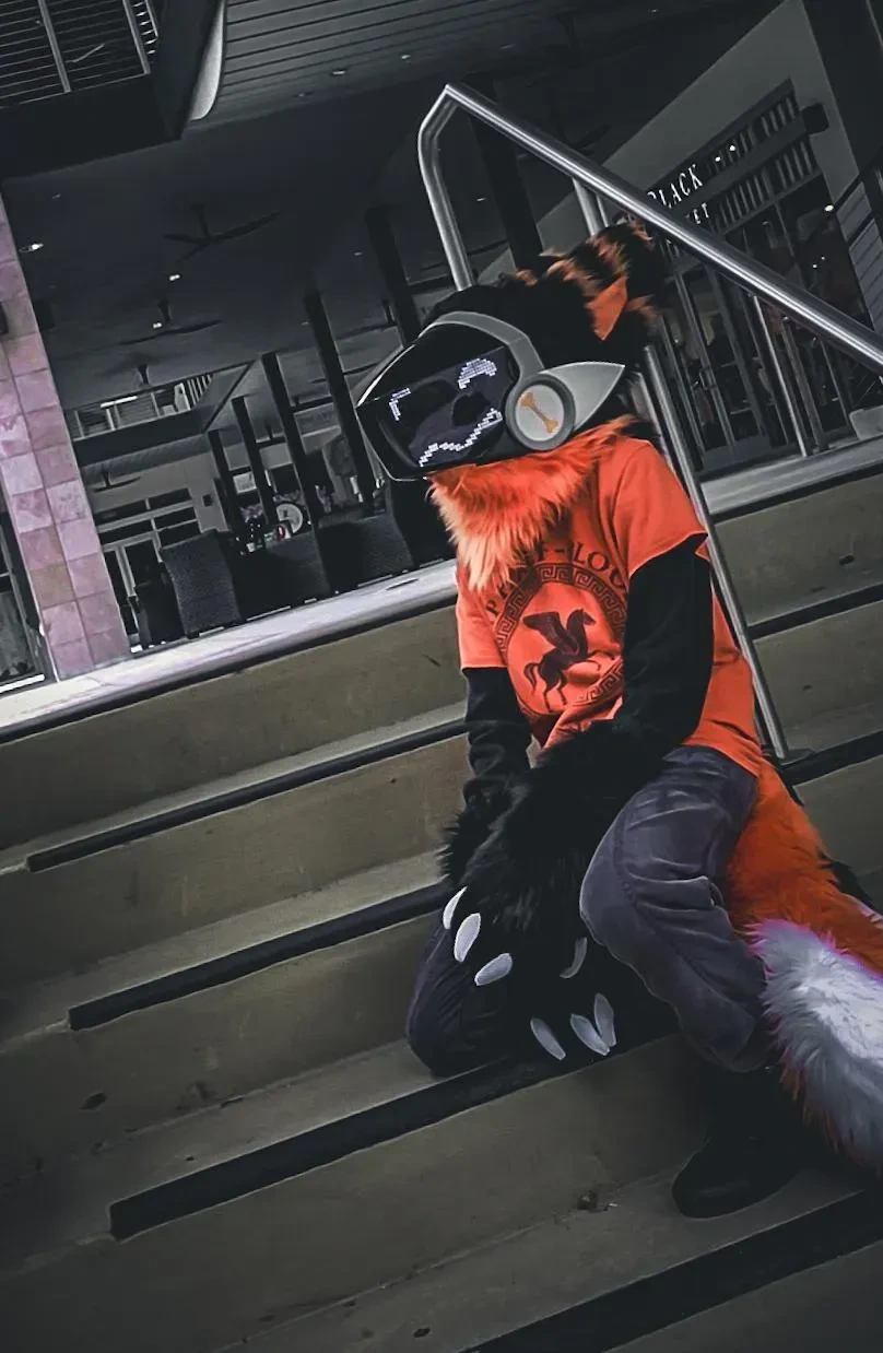 Black and orange fursuit partial with a silver visor and bone emblem, sitting on outdoor steps, by Arget & Lazuli