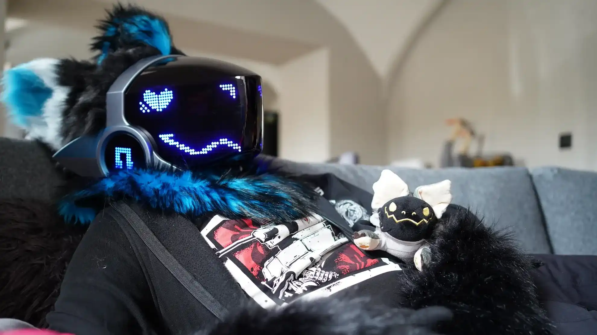 Black and teal Protogen fursuit with an LED heart eyes visor, relaxing on a sofa, by Arget & Lazuli
